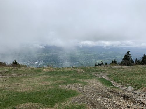 A cloudy view from a mountain top, overlooking a green valley and distant hills.