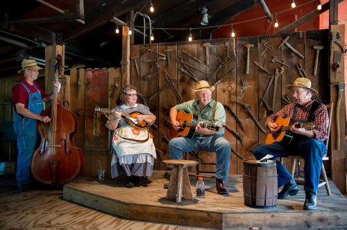 A bluegrass band performs on stage, featuring a bassist, mandolin player, and two guitarists in a rustic setting.