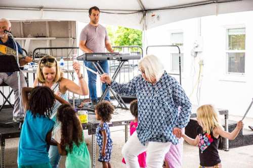 A group of children and adults dance joyfully under a tent, with musicians performing in the background.