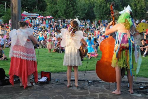 Three young performers in fairy costumes play music on stage, with a vibrant crowd enjoying the outdoor event behind them.