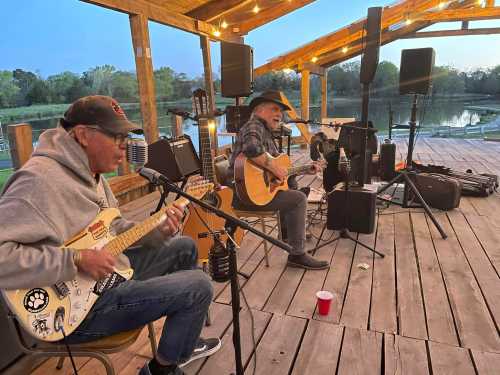 Two musicians perform on a wooden stage by a lake, one playing guitar and the other on an electric guitar.