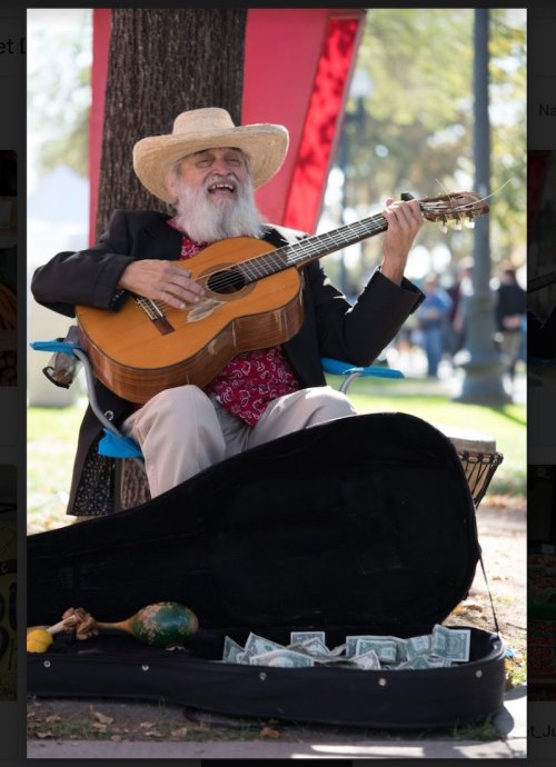 A smiling musician with a guitar sits by a tree, surrounded by money in a guitar case, wearing a straw hat.