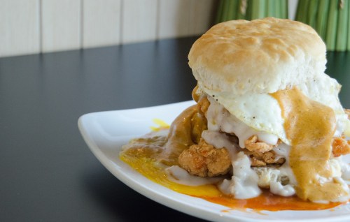 A hearty biscuit sandwich with fried chicken, a fried egg, and gravy, served on a white plate against a dark background.