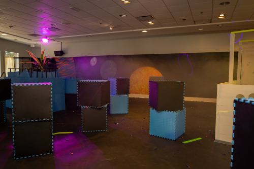 A spacious indoor play area with colorful foam blocks and a mural of planets on the wall, illuminated by purple lights.