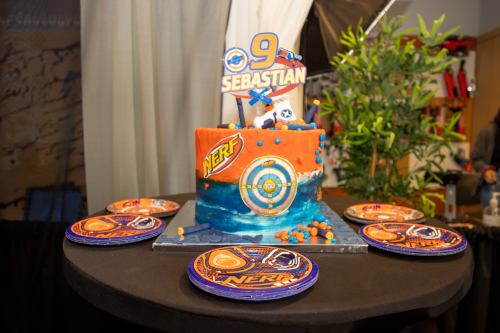 A colorful Nerf-themed birthday cake with blue and orange decorations, featuring a "9" and "Sebastian" on top, surrounded by plates.
