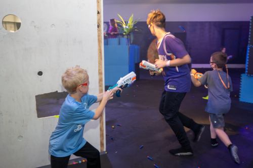 Two children play with toy blasters in a colorful indoor setting, while a third child runs in the background.