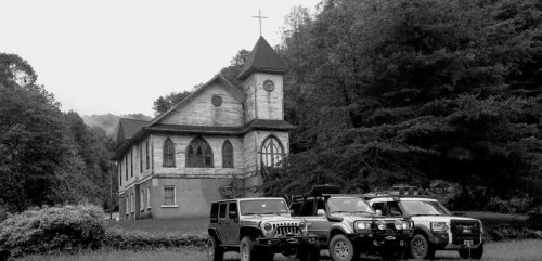 A vintage church with a cross stands behind three off-road vehicles in a rural setting, captured in black and white.