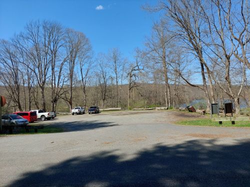 A gravel parking area near a river, surrounded by bare trees and a clear blue sky.