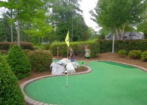 Two children play mini-golf on a green course surrounded by bushes and rocks, with a yellow flag marking the hole.