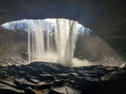 A waterfall cascades through a rocky arch, surrounded by mist and trees under a bright sky.