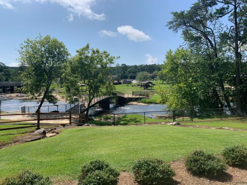 A scenic view of a park with a bridge over a river, green grass, trees, and a clear blue sky.