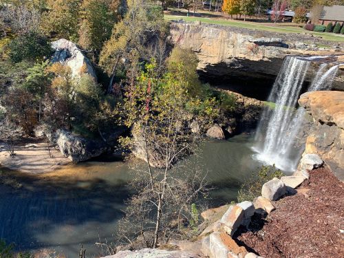 A scenic view of a waterfall cascading into a river, surrounded by rocky cliffs and autumn foliage.