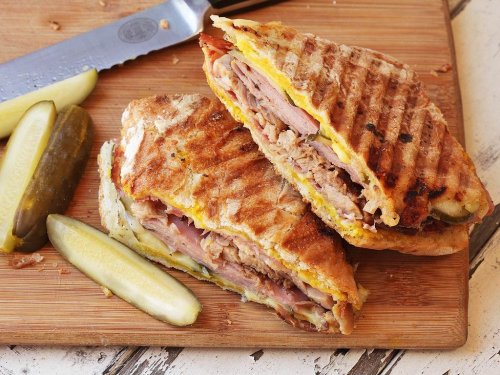 A deliciously grilled sandwich filled with meats, cheese, and pickles, served on a wooden cutting board.