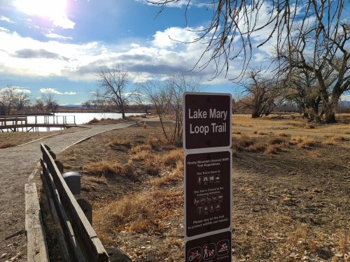 Sign for Lake Mary Loop Trail with a scenic view of a lake and bare trees under a partly cloudy sky.