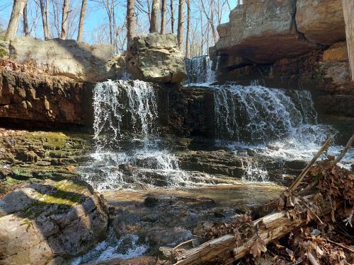 A serene waterfall cascades over rocky ledges, surrounded by bare trees and natural foliage in a peaceful forest setting.