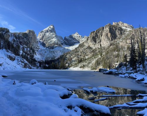 A serene winter landscape featuring a frozen lake surrounded by snow-covered mountains and trees under a clear blue sky.