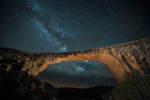 A stunning night sky filled with stars above a large rock arch, illuminated by the Milky Way's glow.