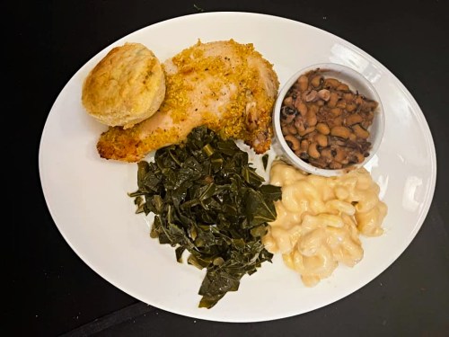 A plate featuring baked chicken, collard greens, macaroni and cheese, black-eyed peas, and a biscuit.