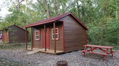 A small wooden cabin with red trim, surrounded by trees, and a picnic table in front on a gravel path.