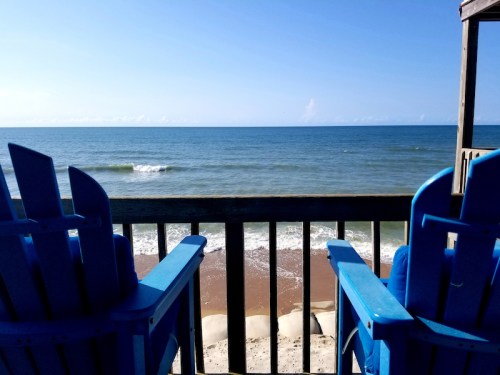 Two blue chairs overlook a calm beach with gentle waves and a clear sky in the background.