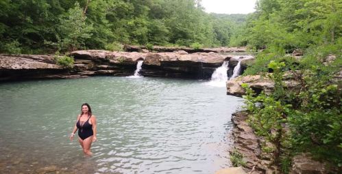 A woman wades in a clear, greenish-blue river surrounded by lush trees and rocky cliffs. Small waterfalls are visible.