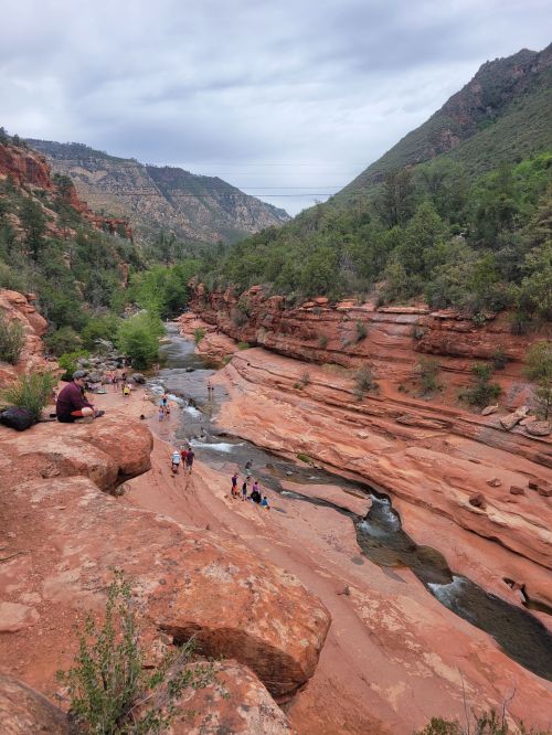 A scenic view of a red rock canyon with a winding river, surrounded by greenery and people enjoying the outdoors.