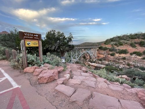 Sign for the Huckaby Trail with a bridge in the background, surrounded by rocky terrain and greenery under a cloudy sky.