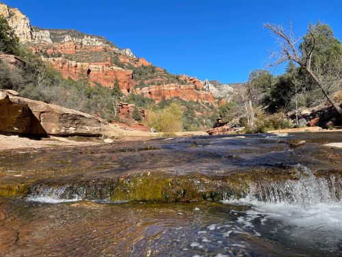 A serene river flows through a rocky landscape, surrounded by red cliffs and green trees under a clear blue sky.