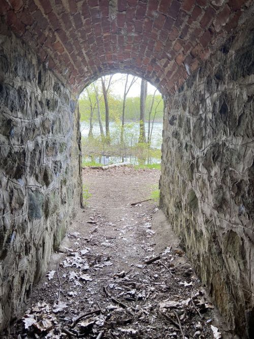 A stone archway leads to a path by a serene body of water, surrounded by trees and greenery.