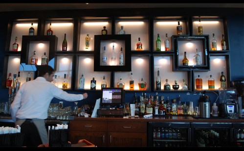 A bartender prepares drinks at a stylish bar with shelves displaying various liquor bottles and a cash register.