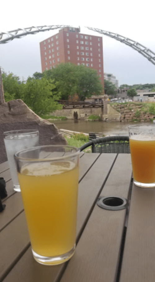 Two glasses of beer on a table with a view of a river and a building in the background under a cloudy sky.