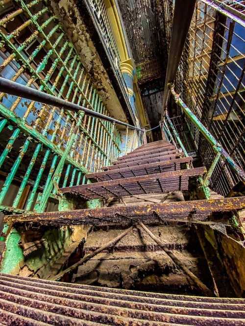 A worn staircase with rusted railings and peeling paint, leading down in a dimly lit, abandoned building.