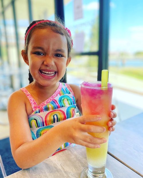 A smiling child holds a colorful layered drink in a bright, sunny café setting.