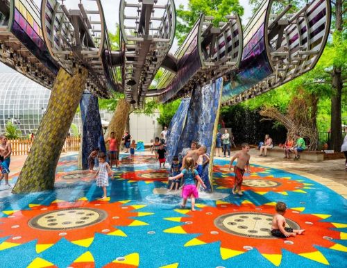 Children and adults play in a colorful splash pad with water features, surrounded by vibrant mosaic patterns and greenery.