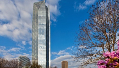 A tall, glass skyscraper under a blue sky with clouds, surrounded by trees and blooming flowers.