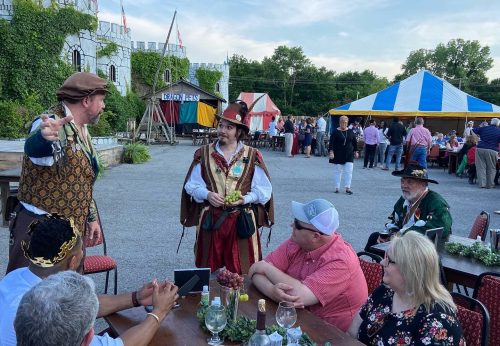 Two performers in period costumes entertain a seated audience at an outdoor event with tents and a castle backdrop.