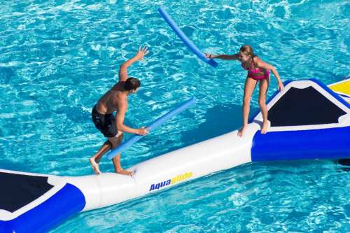 Two children play on a floating inflatable in a pool, using foam noodles for a fun water battle.