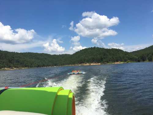 A colorful boat pulls a group on a floating raft across a lake, surrounded by green hills and a blue sky with clouds.