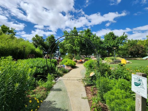 A winding path through a lush garden with greenery, flowers, and a decorative arch under a blue sky with clouds.