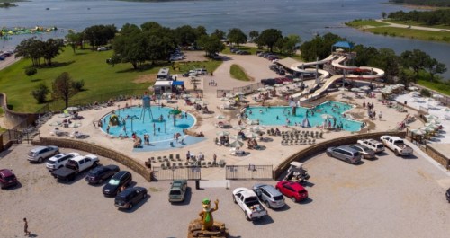 Aerial view of a busy water park with pools, slides, and cars parked nearby, surrounded by trees and a lake.