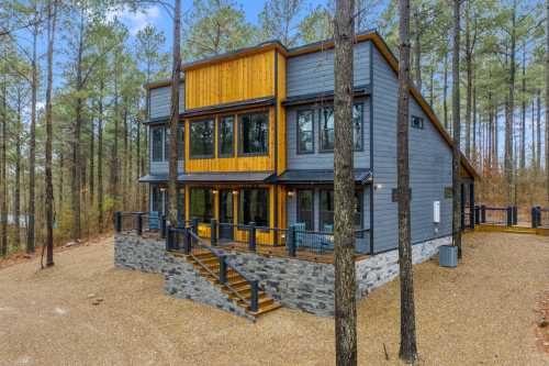 Modern two-story house with large windows, wooden accents, and a stone foundation, surrounded by tall pine trees.