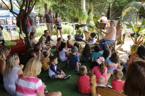 A group of children and adults gathered outdoors, listening to a storyteller among plants and colorful decorations.