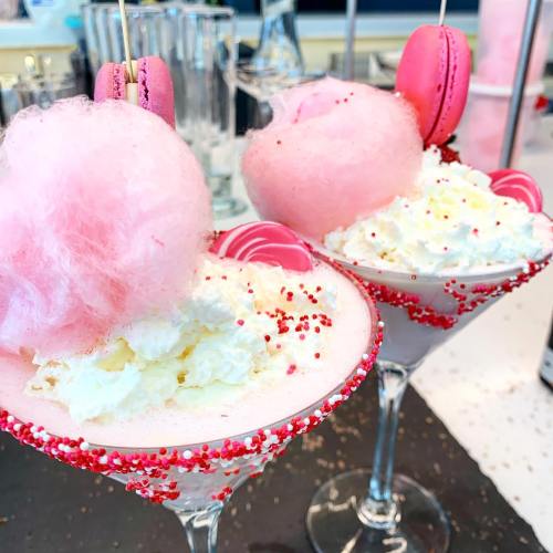 Two colorful cocktails topped with cotton candy, whipped cream, and macarons, garnished with sprinkles.
