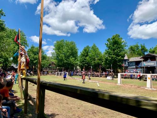 A lively outdoor scene at a festival, featuring performers and a crowd under a bright blue sky with fluffy clouds.