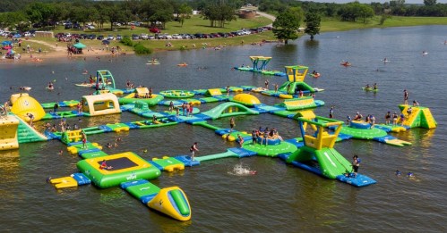 A vibrant inflatable water park with various structures, surrounded by people swimming and enjoying the lake.