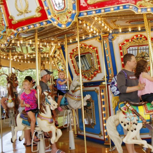 A colorful carousel with children and adults riding on ornate horses, surrounded by bright lights and decorations.