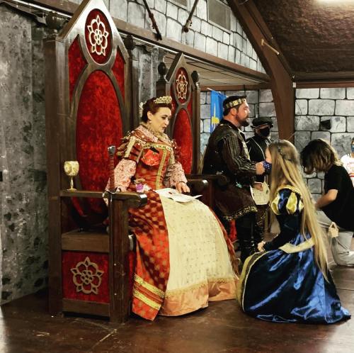 A queen sits on a throne while a girl in a blue dress kneels before her, with a medieval setting in the background.
