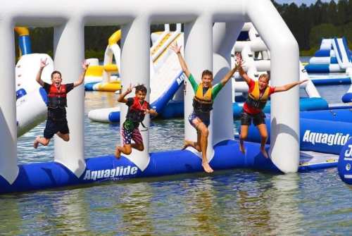 Four children in life jackets joyfully jumping off an inflatable structure into a lake on a sunny day.