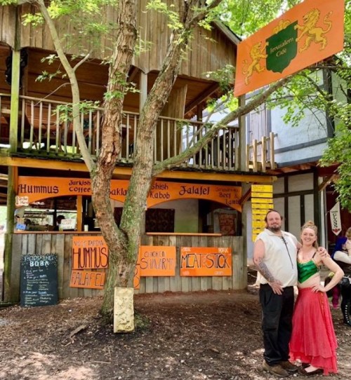A couple poses in front of a rustic café with signs for hummus, falafel, and other dishes, surrounded by trees.