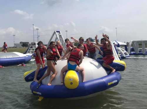 A group of kids in life jackets joyfully poses on a large inflatable water toy in a lake.